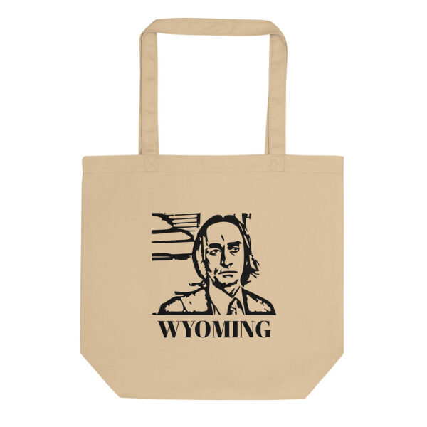 A beige canvas tote bag displays a black stencil-style portrait of a man in front of abstract lines, with the word “WYOMING” in bold capital letters beneath.
