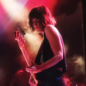 A woman with chin-length dark hair plays bass guitar on stage under dramatic pink, red, and white stage lighting. She wears a sleeveless black top and looks downward with focus.