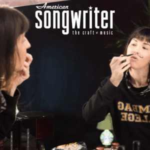 Cover of American Songwriter magazine featuring a woman at a vanity: A woman in a “Scumbag College” sweatshirt draws a curled mustache on her upper lip in a mirror; the reflection is framed by bottles and a glowing lamp. The magazine masthead reads: “American Songwriter – the craft of music.”
