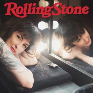 A woman with a tattooed arm rests her head on a towel-covered vanity counter in a backstage dressing room, smiling at the camera. Her reflection appears in a mirror surrounded by glowing globe lights. The word “RollingStone” is written in red at the top.