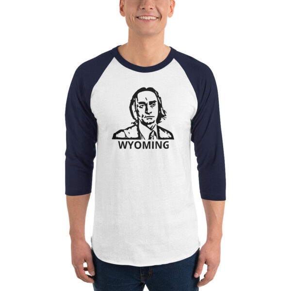 A smiling man wears a white baseball-style shirt with navy blue 3/4 sleeves and a black stencil-style illustration of a man’s face above the word “WYOMING.”