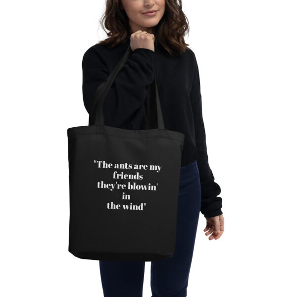 Woman holding a black tote bag with white text that reads: "The ants are my friends they're blowin' in the wind".