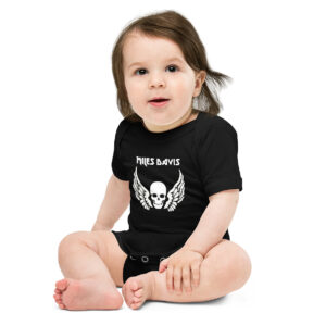Baby in a black onesie featuring a bold white graphic of a skull with wings and the name "MILES DAVIS" in a gothic-style font above it. The baby is sitting upright, legs slightly crossed, with one hand resting on their knee, and has soft brown hair and a gentle smile.