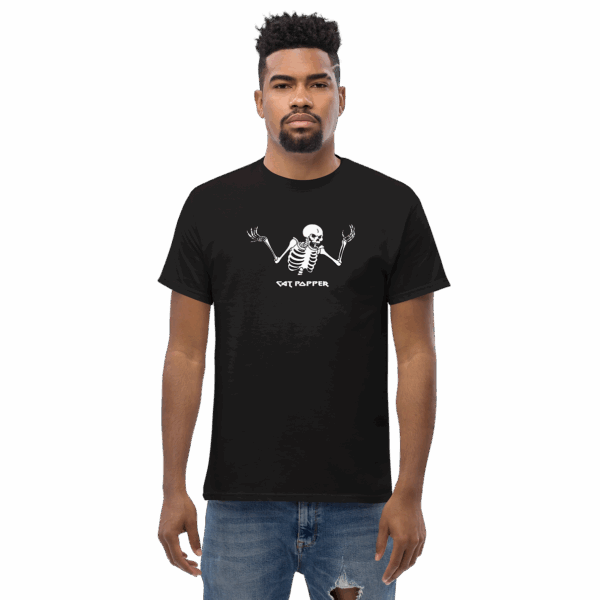 Front-facing view of a young man with short curly hair and a trimmed beard, wearing a black t-shirt with a white skeleton graphic in a shrugging pose and the text "Cat Popper," styled with distressed blue jeans.