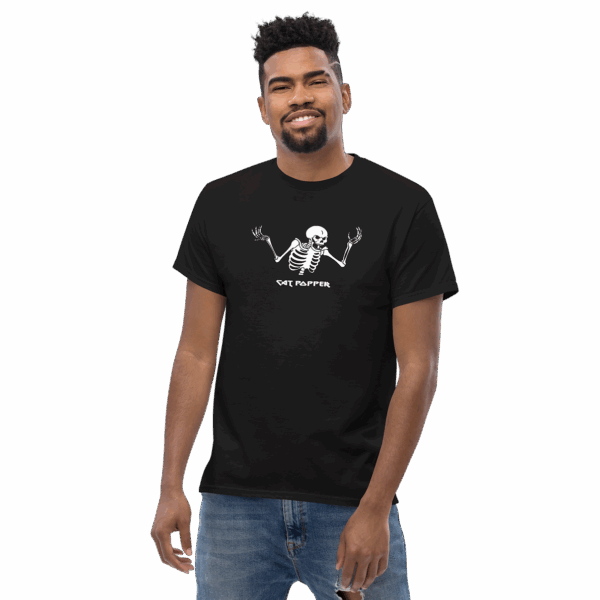 Front view of a young man with short curly hair and a trimmed beard, smiling while wearing a black t-shirt featuring a white graphic of a shrugging skeleton and the text "Cat Popper," paired with ripped blue jeans.