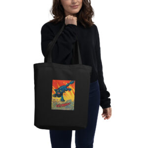 Black canvas tote bag featuring a colorful, comic-style graphic of a fierce blue cat monster leaping through flames with exaggerated claws and teeth. Below the image, the text reads “CAT POPPER” in bold, stylized red letters. A woman in a black sweater and navy pants carries the bag over her shoulder.