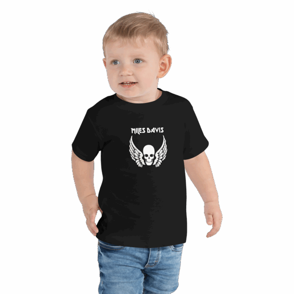 Toddler wearing a black T-shirt with a bold white graphic on the chest that features a skull with outstretched wings beneath the name "MILES DAVIS" in a stylized font. The child has short blond hair, is smiling, and is also wearing denim jeans.