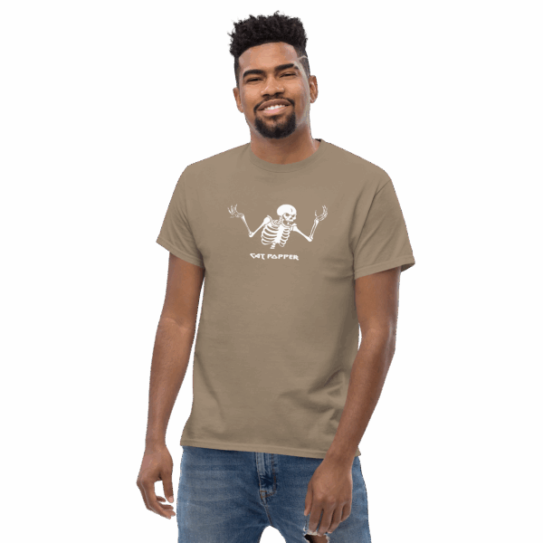 Smiling male model wearing a light brown “Cat Popper” graphic t-shirt featuring a white skeleton illustration with raised arms.