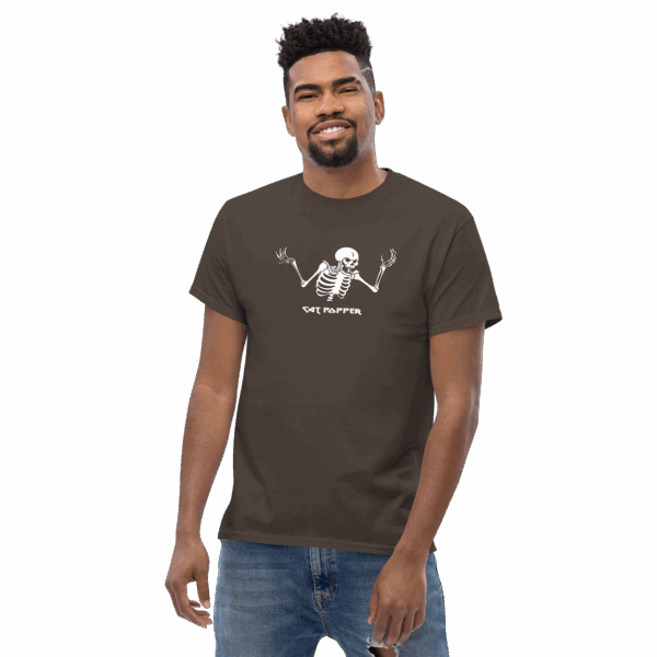 Man wearing a dark brown graphic t-shirt with a white cartoon skeleton illustration and the text “Cat Popper”; he is smiling and standing casually with one hand by his side.