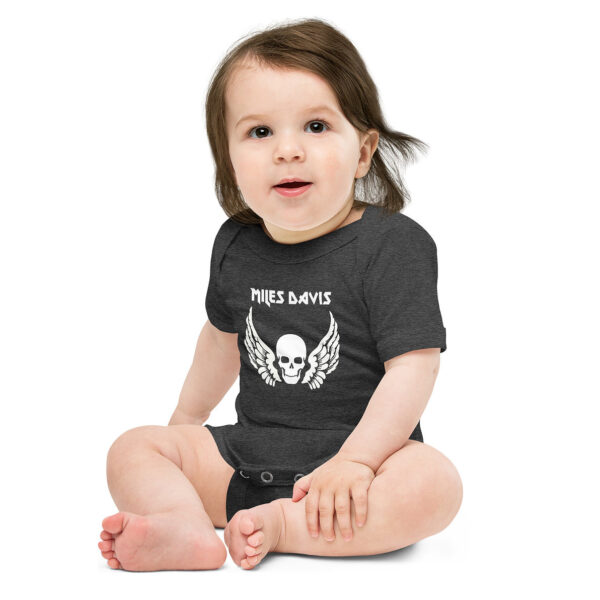 Baby wearing a dark charcoal gray onesie with a white graphic featuring a skull with wings underneath the name "MILES DAVIS" in a bold, gothic-style font. The baby is sitting upright with legs crossed, smiling softly, and has medium-length brown hair with a playful, slightly tousled look.