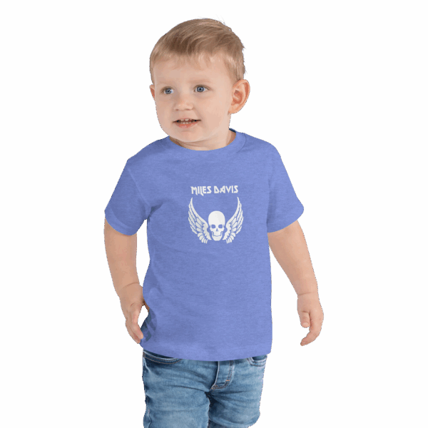 Toddler in a heather blue T-shirt featuring a white skull and wings graphic with the name "MILES DAVIS" in a bold, stylized font above the image. The child is smiling, with one hand in their pocket, and wearing denim jeans.