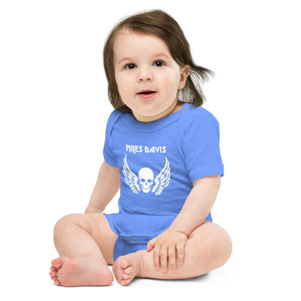 Baby wearing a bright blue onesie featuring a bold white graphic of a skull with angel wings beneath the name "MILES DAVIS" in a sharp, rock-inspired font. The baby is seated cross-legged with a gentle expression, slightly smiling, and has tousled brown hair falling over one eye.