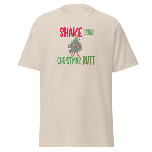 Light beige short-sleeve t-shirt featuring a festive design with the phrase "Shake Your Christmas Butt" in bold red, green, and brown text, centered above and below a cartoon Christmas tree character shaking its hips playfully.