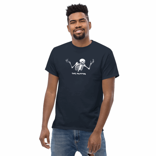 A smiling man wearing a navy blue T-shirt featuring a white skeleton graphic with raised arms and the text “Cat Popper” beneath it, standing casually with one arm relaxed.