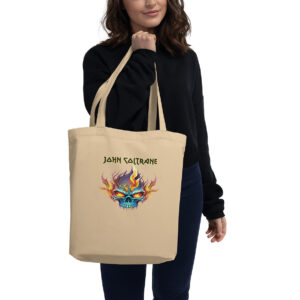 Beige canvas tote bag with a graphic of a flaming blue skull in vivid colors and the name “John Coltrane” above it in bold, stylized black lettering. A woman in a black sweater and dark jeans is holding the bag over her shoulder.