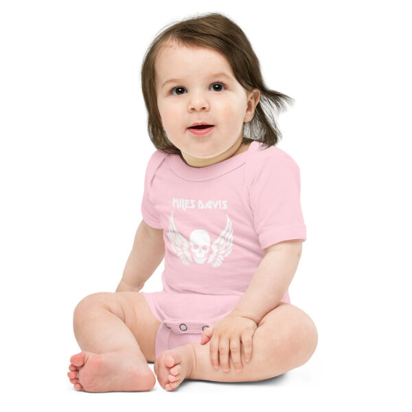 Baby wearing a light pink onesie featuring a white graphic of a winged skull with the text "MILES DAVIS" above it in a bold, angular font. The baby is sitting on the floor, legs bent and crossed, one hand resting on their foot and the other on their leg, with a soft, curious expression and dark hair falling gently to the side.