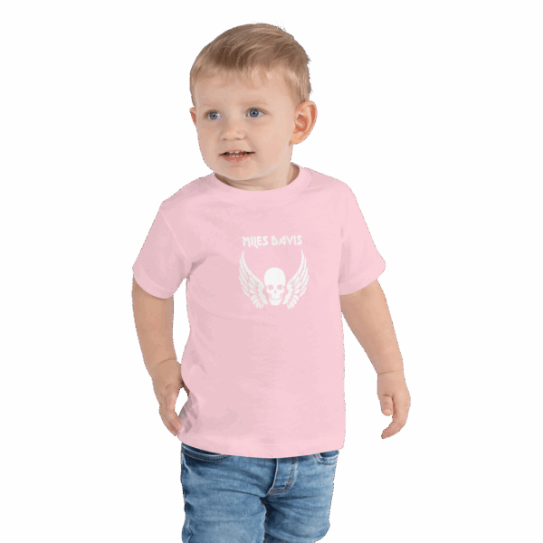 Toddler in a light pink T-shirt featuring a white graphic design of a skull with angel wings and the name "MILES DAVIS" in a bold, stylized font above it. The child is standing, smiling slightly, and wearing denim jeans with their right hand partially in their pocket.