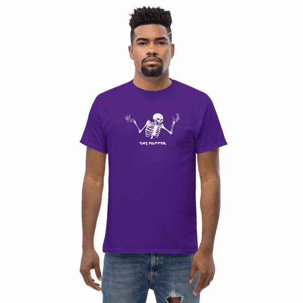 A man wearing a bold purple T-shirt featuring a central white graphic of a skeleton raising its arms in a dramatic shrug. The skeleton’s head is tilted, with expressive hands up and fingers splayed. Below the skeleton, the name “CAT POPPER” is written in a stylized, playful font. The man stands facing forward with a neutral expression, wearing light distressed jeans.