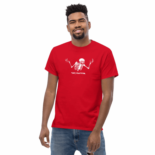 A man in a red T-shirt with a white skeleton graphic on the chest. The skeleton has both arms raised in a shrugging pose, and the text below reads “CAT POPPER” in a bold, playful font. The man is smiling slightly and standing casually with one arm relaxed and the other by his side. He's wearing light-wash jeans with a rip in the knee.