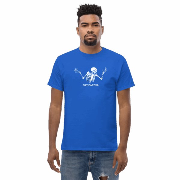 A man standing straight, wearing a royal blue T-shirt with a white skeleton graphic on the chest. The skeleton has both arms raised in a shrugging pose, and below it, the text reads “CAT POPPER” in a bold, playful font. The man has a serious facial expression and is wearing distressed jeans.